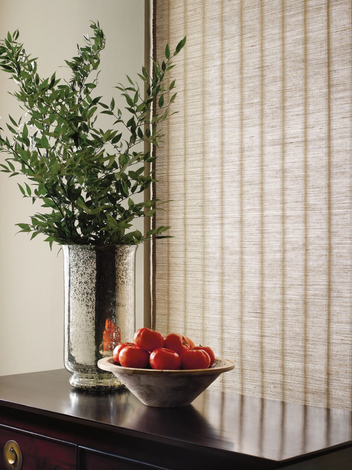 a vase and a bowl of fruit in front of woven wood shades