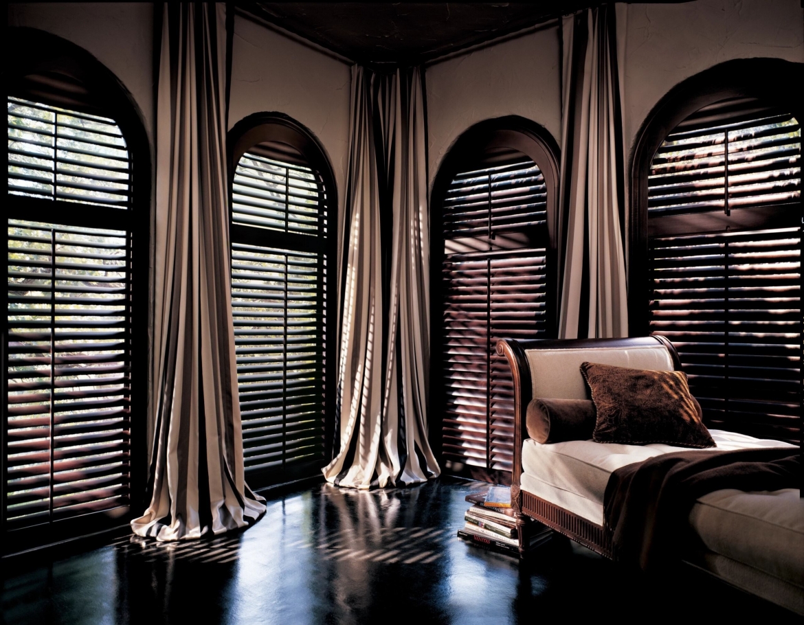 A darkly shaded room with an easy chair and plantation shutters on the windows